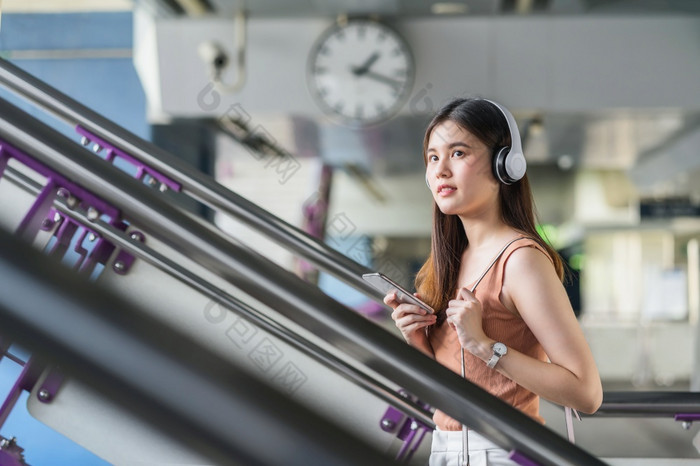 年轻的亚洲女人乘客穿耳机和使用聪明的移动电话听音乐和走的楼梯地铁站当旅行大城市通勤和运输概念
