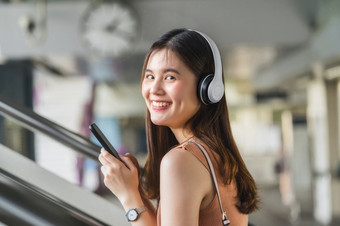 年轻的亚洲女人乘客穿耳机和使用聪明的移动电话听音乐和走的楼梯地铁站当旅行大城市通勤和运输概念