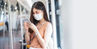 年轻的亚洲女人乘客穿外科手术面具和听音乐通过聪明的移动电话地铁火车当旅行大城市科维德爆发感染和流感大流行概念