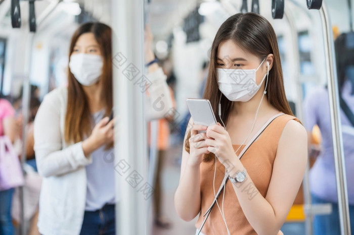 年轻的亚洲女人乘客穿外科手术面具和听音乐通过聪明的移动电话地铁火车当旅行大城市科维德爆发感染和流感大流行概念