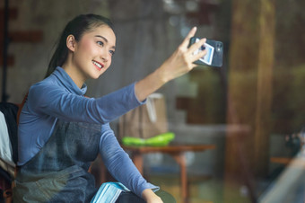 亚洲年轻的快乐的女人使自我肖像使用聪明的移动电话咖啡商店酒吧俱乐部有吸引力的微笑小业务老板与围裙<strong>照片</strong>为她的<strong>社会</strong>网络