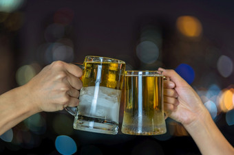 手持有和无比的与两个啤酒杯子眼镜在的照片模糊城市景观为庆祝聚会，派对和庆祝与快乐新一年概念