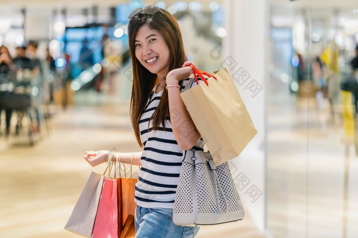 快乐亚洲女人持有的购物袋和看为新时尚衣服通过玻璃商店商店