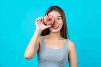 有吸引力的亚洲年轻的女人微笑和玩与甜蜜的甜甜圈她的脸孤立的蓝色的颜色背景重量损失和避免垃圾食物为节食和健康的概念复制空间和工作室