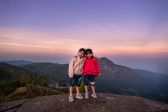 两个可爱的小女孩拥抱前山和看的相机对日落背景徒步旅行冒险与家庭假期