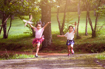 快乐孩子们玩与纸板飞机的夏天花园两个小姐妹与玩具飞机运行的路的公园快乐童年概念