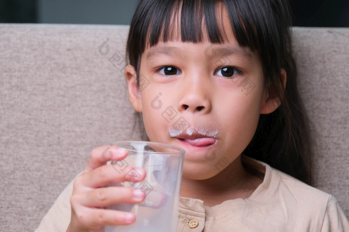 肖像可爱的亚洲小女孩持有玻璃牛奶坐着的沙发首页小女孩首页与微笑脸感觉快乐享受喝牛奶和看相机