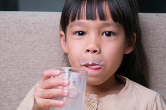 肖像可爱的亚洲小女孩持有玻璃牛奶坐着的沙发<strong>首页</strong>小女孩<strong>首页</strong>与微笑脸感觉快乐享受喝牛奶和看相机