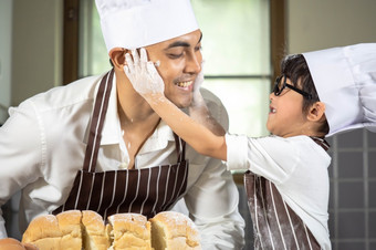 亚洲男孩穿眼镜取笑爸爸烹饪与白色面粉揉捏面包面团教孩子们实践烘焙成分面包蛋餐具厨房生活方式快乐学习生活与家庭