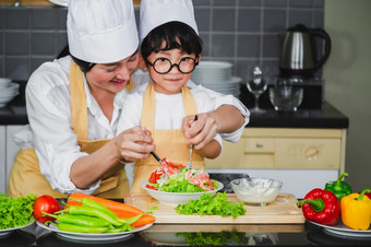 亚洲女人年轻的妈妈。与儿子男孩烹饪沙拉妈妈切片蔬菜食物儿子品尝沙拉沙拉酱蔬菜胡萝卜和西红柿贝尔辣椒快乐家庭烹饪食物享受生活方式厨房
