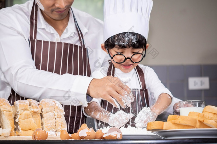亚洲男孩穿眼镜取笑爸爸烹饪与白色面粉揉捏面包面团教孩子们实践烘焙成分面包蛋餐具厨房生活方式快乐学习生活与家庭有趣的学习