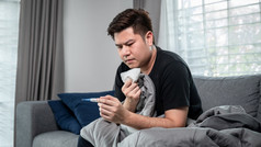 亚洲男人。使用组织咳嗽打喷嚏和使用温度计检查身体温度当感觉生病的和坐着与毯子的沙发上