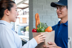亚洲交付男人。蓝色的统一的持有杂货店袋食物水果蔬菜给女人客户前面房子那服务在冠状病毒新冠病毒流感大流行