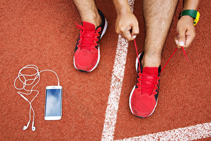 跑步者年轻的男人。系鞋带运行鞋子鞋带得到准备好了为比赛运行跟踪与智能手机和耳机为音乐听移动电话运动员准备为有氧运动培训