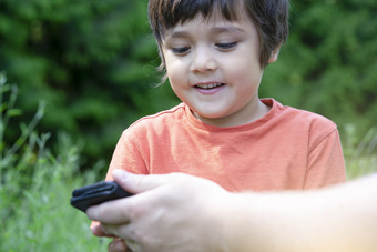 裁剪拍摄孩子男孩选址的公园与父玩游戏聪明的电话孩子看移动电话与模糊的绿色自然背景快乐家庭概念