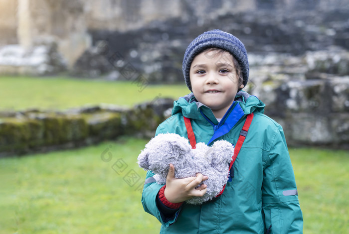 肖像学校孩子采取泰迪熊探索与他的学习历史快乐孩子男孩穿温暖的布持有他的软玩具站独自一人与模糊的废墟老修道院背景