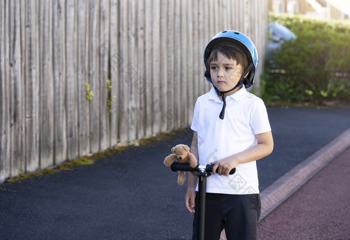 肖像学校男孩骑踏板车学校孩子穿安全头盔骑辊孩子站与泰迪熊看深inthought活跃的休闲和户外体育运动为孩子们