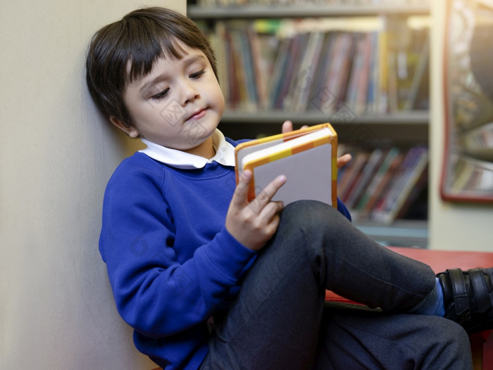 学前教育孩子坐着红色的沙发享受阅读卡通的图书馆的学校可爱的男孩与兴奋脸而阅读最喜欢的书图书馆早期一年学生活动概念