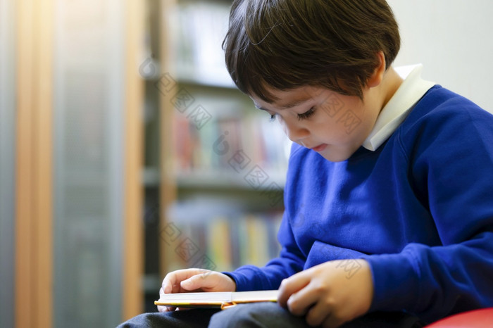学前教育孩子sittng红色的沙发享受阅读卡通的图书馆的学校可爱的男孩与兴奋脸而阅读最喜欢的书图书馆早期一年学生活动概念