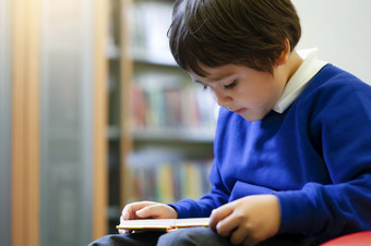 学前教育孩子sittng红色的沙发享受阅读卡通的图书馆的学校可爱的男孩与兴奋脸而阅读最喜欢的书图书馆早期一年学生活动概念