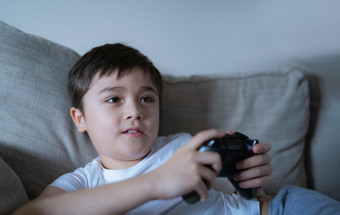 快乐年轻的男孩玩视频游戏在线与朋友坦诚的拍摄可爱的孩子坐着沙发持有游戏控制台肖像孩子看监控而玩游戏和放松首页