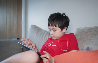 孩子坐着沙发看卡通玩游戏平板电脑孩子男孩使用数字垫学习教训在线互联网首页学校教育距离学习在线教育概念