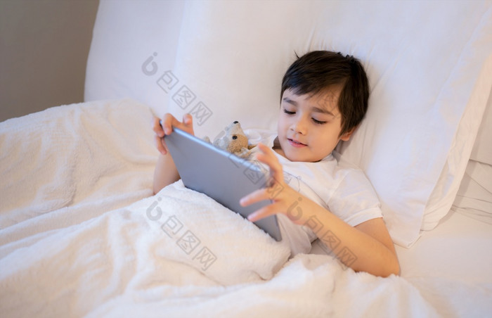 快乐孩子说谎床上持有平板电脑看卡通和聊天与朋友数字垫可爱的年轻的男孩玩游戏在线互联网孩子放松的早....之前学校