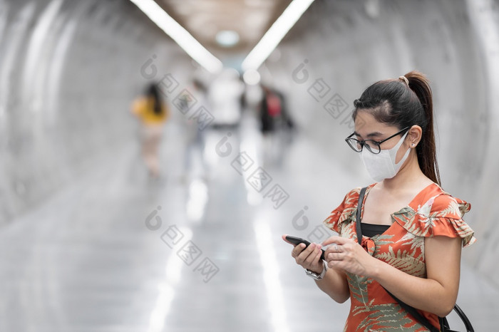 年轻的亚洲女孩穿外科手术脸面具保护冠状病毒变形快乐旅游女人使用智能手机在走地铁站新正常的和生活后新冠病毒流感大流行