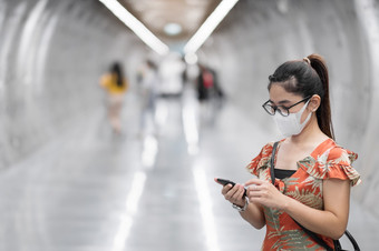 年轻的亚洲女孩穿外科手术脸面具保护冠状病毒变形快乐旅游女人使用智能手机在走地铁站新正常的和生活后新冠病毒流感大流行