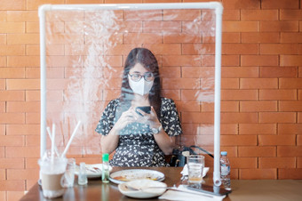 亚洲女人使用智能手机在餐厅时间餐厅与分离盾塑料分区保护冠状病毒变形社会距离新正常的和生活后新冠病毒流感大流行