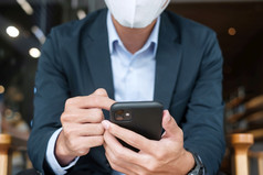 年轻的商人西装穿外科手术脸面具和使用智能手机男人。打字触屏移动电话办公室咖啡馆新冠病毒流感大流行技术和新正常的概念