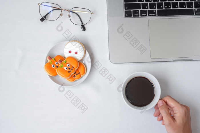 手持有咖啡杯和吃万圣节饼干在使用电脑移动PC快乐万圣节在线购物你好10月秋天秋天节日聚会，派对和假期概念