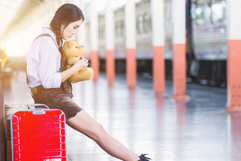 亚洲怀孕了女人旅行者坐着拥抱泰迪熊与红色的手提箱铁路站旅行肚子女孩