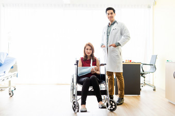 亚洲年轻的男人。医生检查夹板的手臂女病人手由于与她的手臂破碎的为更好的疗愈与微笑坐轮椅的房间医院背景
