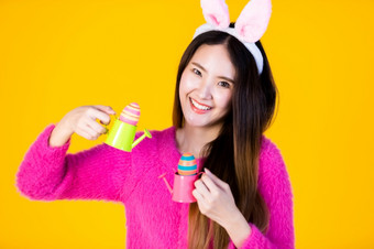 复活节假期概念快乐微笑亚洲年轻的女人穿<strong>兔子</strong>耳朵持有与小浇水可以色彩斑斓的复活节鸡蛋和的一边<strong>眼睛</strong>看相机孤立的黄色的空白复制