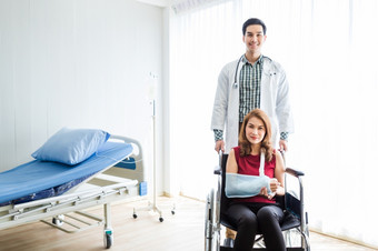 亚洲年轻的男人。医生检查夹板的手臂女病人手由于与她的手臂破碎的为更好的疗愈与微笑坐轮椅的房间<strong>医院背景</strong>