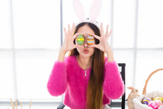 快乐微笑亚洲年轻的女人穿兔子耳朵和持有色彩斑斓的复活节蛋前面她的眼睛串的白色房间背景