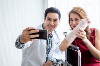 自拍微笑男人。医生与女病人穿手臂夹板为更好的疗愈坐轮椅使用智能手机的房间医院背景