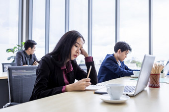 心烦意乱深思熟虑的年轻的亚洲业务女人压力的工作场所工作与移动PC电脑办公室工作人员是不快乐与工作的同事们后面的办公室