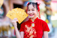 肖像美丽的微笑可爱的小亚洲女孩穿红色的传统的中国人旗袍装饰持有黄色的信封手为中国人新一年节日中国人神社