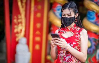 亚洲年轻的女人穿红色的传统的<strong>中国</strong>人旗袍装饰和穿保护面具和智能<strong>手机</strong>为<strong>中国</strong>人新一年节<strong>日</strong>神社预防的传播新冠病毒病毒