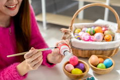 复活节假期概念快乐亚洲年轻的女人穿兔子耳朵手画鸡蛋为复活节与色彩斑斓的复活节鸡蛋的白色房间背景