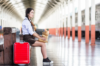 亚洲怀孕了女人旅行者坐着拥抱泰迪熊与红色的手提箱铁路站旅行旅行者与背包夏天假期概念泰国