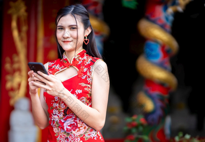 肖像美丽的微笑亚洲年轻的女人穿红色的旗袍衣服传统的装饰和写作消息智能手机为中国人新一年节日中国人神社泰国
