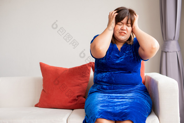 超重亚洲女人坐着的沙发的生活房间和持有手的头由于头痛从偏头痛概念疾病和健康哪