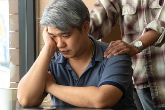 中年亚洲男人。年老强调和累了是坐着快食物餐厅和有朋友站后面鼓励概念帮助和鼓励