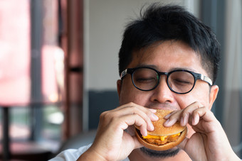 亚洲男人。吃汉堡快食物餐厅和享受美味的食物男人。白色t恤和眼镜持有汉堡和享受的味道汉堡