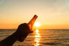 手男人。持有啤酒瓶和持有他的手的天空晚上与日落庆祝假期的海滩夏天