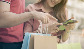 快乐年轻的夫妇购物者走的购物街对和持有色彩斑斓的购物袋手和使用智能手机为检查促销活动概念出售和黑色的星期五购物