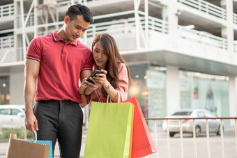 快乐年轻的夫妇购物者走的购物街对和持有色彩斑斓的购物袋手和使用智能手机为检查促销活动概念出售和黑色的星期五购物